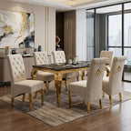 A 7-piece dining set featuring a rectangular black marble-top table with gold legs, surrounded by six tufted cream upholstered dining chairs with gold legs, set in a modern dining room.
