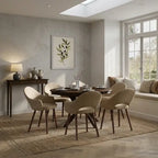 A dining room scene featuring a round dark wood table surrounded by six upholstered dining chairs in a warm beige fabric, set on a woven area rug. The room has a textured light gray wall, a built-in window seat with cushions, and a framed botanical print on the wall.