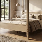 A light natural wood four-poster bed frame styled with neutral beige and taupe bedding, set in a bright, modern bedroom 