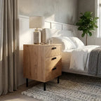 A light oak three-drawer nightstand with black metal legs and dark handles, styled in a modern bedroom next to a bed with white linens and a bedside lamp.