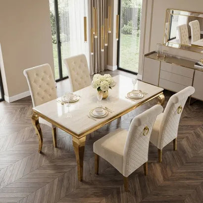 A luxury dining room set featuring a rectangular table with a white marble-look top and ornate gold legs, surrounded by four tufted cream upholstered chairs with gold accents, set in a modern room with herringbone wood flooring.