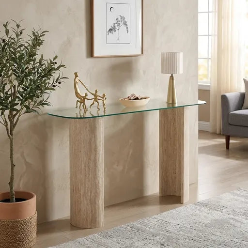 A modern console table with a clear glass top and two textured light beige stone legs, styled in a warm, 