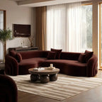 A modern curved sectional sofa upholstered in deep burgundy velvet, styled in a luxurious living room with a round wooden coffee table, a striped beige and white rug, and sheer white curtains.