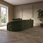A modern curved sofa upholstered in dark olive green velvet, styled on a textured beige rug in a minimalist room with beige paneled walls, light wood herringbone flooring, and a potted plant.