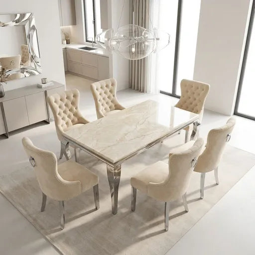A modern dining room set featuring a rectangular marble-top dining table with a polished silver base, surrounded by six tufted cream velvet chairs with silver legs, set on a light beige rug in a bright contemporary room.