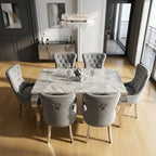 A modern dining set featuring a rectangular marble-patterned table and six tufted gray velvet chairs with silver nailhead trim, shown in a bright, contemporary dining room.