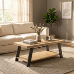 A modern wooden coffee table with a light oak top and dark gray angled legs, styled in a bright living room next to a cream sofa and a potted plant.