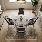 A round marble dining table set with four gray tufted velvet chairs, placed on a large patterned area rug in a bright, modern dining room with light wood floors.