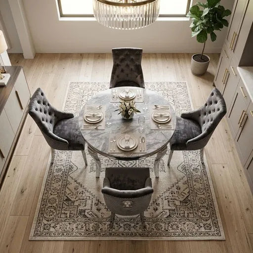 A round marble dining table set with four gray tufted velvet chairs, placed on a large patterned area rug in a bright, modern dining room with light wood floors.
