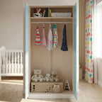 A tall children's wardrobe with light blue doors and a beige interior, shown open in a nursery. The wardrobe contains several colorful dresses hanging on hangers, a storage box with stuffed teddy bears, and a small wooden toy on the bottom shelf.