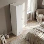 A tall, minimalist white storage cabinet with a single door and a small silver handle, shown in a modern, sunlit bedroom next to a bed and chair.