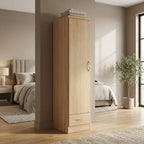 A tall, narrow wardrobe or storage cabinet in a light natural wood finish stands in a modern bedroom setting. The cabinet features a single door with a round metallic handle and a small bottom drawer with a silver pull, with folded towels stacked on top.
