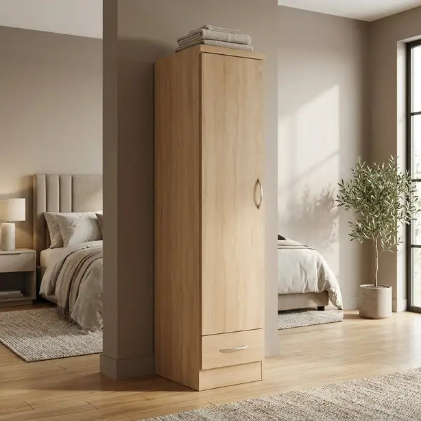 A tall, narrow wardrobe or storage cabinet in a light natural wood finish stands in a modern bedroom setting. The cabinet features a single door with a round metallic handle and a small bottom drawer with a silver pull, with folded towels stacked on top.