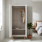 A tall white two-door wardrobe with light wood legs stands open in a bright bedroom. The interior has a light gray wood-look back panel, a single shelf, and two wooden hangers with hanging garments.