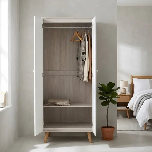 A tall white two-door wardrobe with light wood legs stands open in a bright bedroom. The interior has a light gray wood-look back panel, a single shelf, and two wooden hangers with hanging garments.