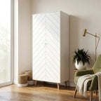 A tall white  two-door wardrobe with a chevron pattern on the doors and tapered wooden legs, styled in a bright room next to a green armchair and a window.