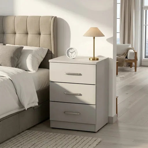 A three-drawer nightstand in a light gray finish with silver handles, styled next to a bed in a modern bedroom. A small table lamp and a round clock sit on top of the nightstand.
