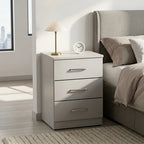 A three-drawer nightstand in a light gray finish, styled next to a bed in a modern bedroom. The nightstand features silver metal drawer pulls and sits next to a brass table lamp and a small white clock.