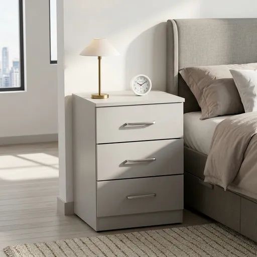 A three-drawer nightstand in a light gray finish, styled next to a bed in a modern bedroom. The nightstand features silver metal drawer pulls and sits next to a brass table lamp and a small white clock.