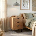 A three-drawer wooden nightstand with a geometric drawer front and black metal legs, styled next to a bed in a softly lit bedroom.