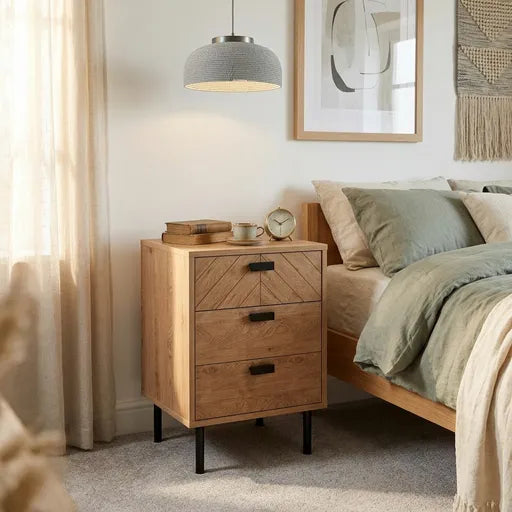 A three-drawer wooden nightstand with a geometric drawer front and black metal legs, styled next to a bed in a softly lit bedroom.