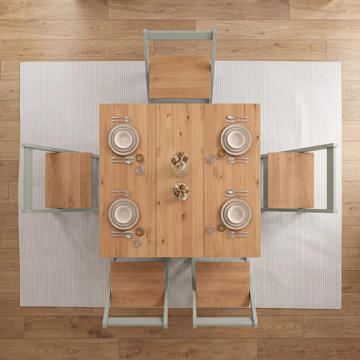 Overhead view of a rectangular wooden dining table set with four chairs, white plates, silverware, and a small glass vase, placed on a white ribbed rug over a wood floor.