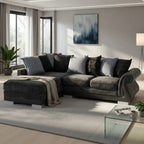 A modern L-shaped sectional sofa in dark gray velvet fabric, styled with multiple gray and black throw pillows and a beige knit blanket, set in a contemporary living room with a light gray rug and glass coffee table.