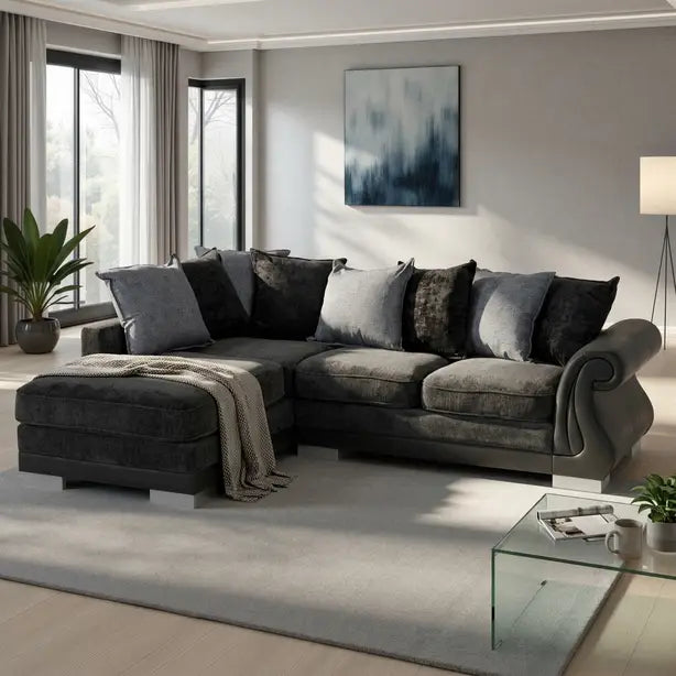 A modern L-shaped sectional sofa in dark gray velvet fabric, styled with multiple gray and black throw pillows and a beige knit blanket, set in a contemporary living room with a light gray rug and glass coffee table.