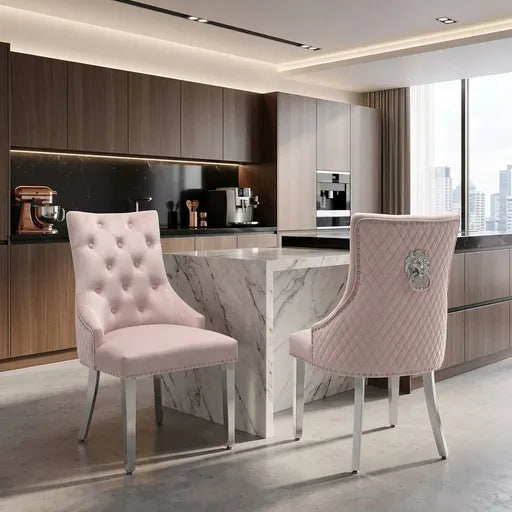 Two elegant blush pink upholstered dining chairs with silver metal legs, displayed in a modern kitchen setting with a marble island and dark wood cabinetry.