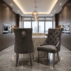 Two modern gray upholstered dining chairs in a luxury kitchen setting, one with a quilted diamond pattern and lion head knocker, the other with tufted velvet upholstery.