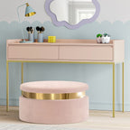 Pink vanity table with gold legs and a matching pink ottoman with gold band, against a light blue wall.