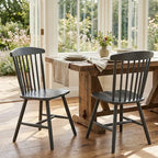 Two dark gray spindle-back dining chairs positioned on a rustic wooden table in a bright dining room with large windows and a garden view.