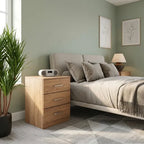 A modern bedroom scene featuring a light oak three-drawer nightstand next to a bed with beige bedding and gray pillows. The room has sage green walls, a gray geometric rug, and a potted green plant in the foreground.
