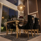 A luxury dining room set featuring a rectangular black dining table with polished gold legs, surrounded by four black tufted velvet chairs with gold lion head accents and nailhead trim. The scene is illuminated by a geometric gold pendant light and warm ambient lighting from a modern interior with wood paneling and large windows.