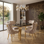 A luxury dining room set featuring a rectangular black marble-top table with a gold frame, surrounded by four tufted blush pink velvet chairs with gold accents. The room is styled with a modern chandelier, wood herringbone flooring, and a dark wood accent wall with gold geometric lines.