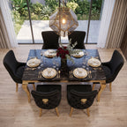 A luxury dining room setup featuring a rectangular black marble dining table with gold legs, surrounded by six black tufted velvet chairs with gold lion-head handles. The table is set with gold-rimmed white plates, gold cutlery, and wine glasses, with a central floral arrangement.