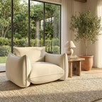 Beige armchair in a room with large windows, a wooden side table, and a potted plant.
