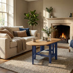 A blue wooden coffee table with a light wood oval top and lower shelf, styled in a cozy living room next to a beige sofa and a burning fireplace.