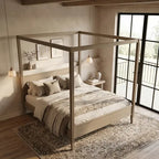 A light wood four-poster canopy bed with neutral bedding, styled in a warm, modern minimalist bedroom with a patterned rug, bedside tables, and large black-framed windows.
