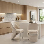 A modern kitchen interior with light wood cabinetry, white countertops, and two upholstered dining chairs in the foreground. The chairs have a textured cream fabric and curved sculptural backs with slim metallic legs.