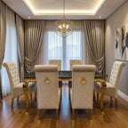 A luxury dining room featuring a rectangular dining table with a dark marble top and gold legs, surrounded by six high-back tufted cream chairs with gold ring pulls. The room is decorated with elegant draped curtains, a geometric gold chandelier, and hardwood flooring.