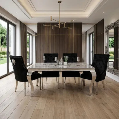 A modern dining room featuring a rectangular white marble dining table with six black tufted velvet chairs, set in a spacious room with light wood floors, a dark wood accent wall, and a gold chandelier.