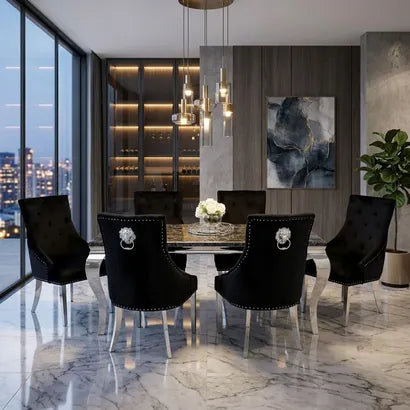 A modern dining room scene with a rectangular glass-top dining table surrounded by six black upholstered chairs with silver metal legs and decorative lion-head ring pulls. The room has marble flooring, a dark wood wall, built-in shelving, pendant lights, and a large abstract painting.