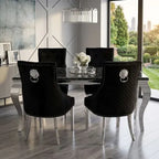 A luxury dining room set featuring a black glass-top table surrounded by four black upholstered dining chairs with silver lion head ring pulls and silver nailhead trim, set in a modern room with a large window overlooking a green garden.