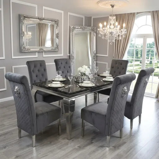 A luxury dining room set featuring a rectangular black marble-top table with a mirrored glass base, surrounded by six gray tufted velvet high-back chairs with silver metal legs. The room is styled with elegant silver-framed mirrors, a crystal chandelier, and gray paneled walls.