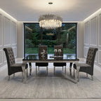 A luxury dining room set featuring a rectangular black marble-top table with silver legs, surrounded by four tufted gray velvet chairs. The room is decorated with a large modern chandelier, white paneled walls, and a large window overlooking a green garden.