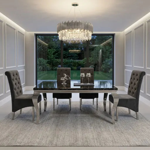 A luxury dining room set featuring a rectangular black marble-top table with silver legs, surrounded by four tufted gray velvet chairs. The room is decorated with a large modern chandelier, white paneled walls, and a large window overlooking a green garden.