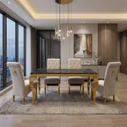 A modern dining room set featuring a rectangular dark marble-top table with ornate gold legs, surrounded by four tufted cream upholstered dining chairs and two matching stools, set on a patterned area rug in a contemporary loft.