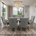 A luxury dining room set featuring a rectangular distressed wood table surrounded by six tufted gray velvet chairs with silver nailhead trim and ring pulls, set on a patterned area rug under a large crystal chandelier.