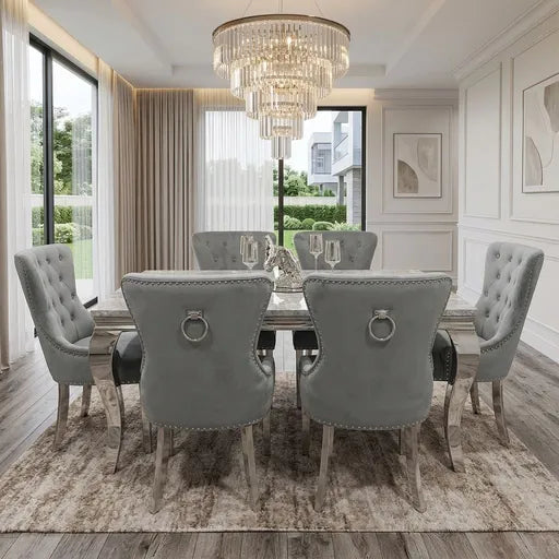 A luxury dining room set featuring a rectangular distressed wood table surrounded by six tufted gray velvet chairs with silver nailhead trim and ring pulls, set on a patterned area rug under a large crystal chandelier.